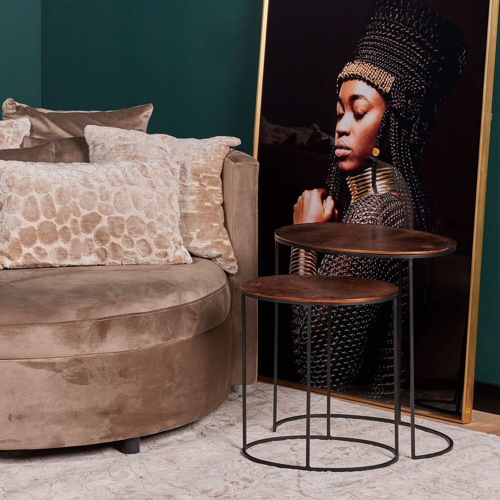 Goedkope ronde bijzettafels van koperen blad en zwart metalen poten in een trendy modern interieur met donkere muren en een zwart met goud schilderij tegen de muur aan geleund, en een zachte roze stoel in de hoek met sierkussens er op.